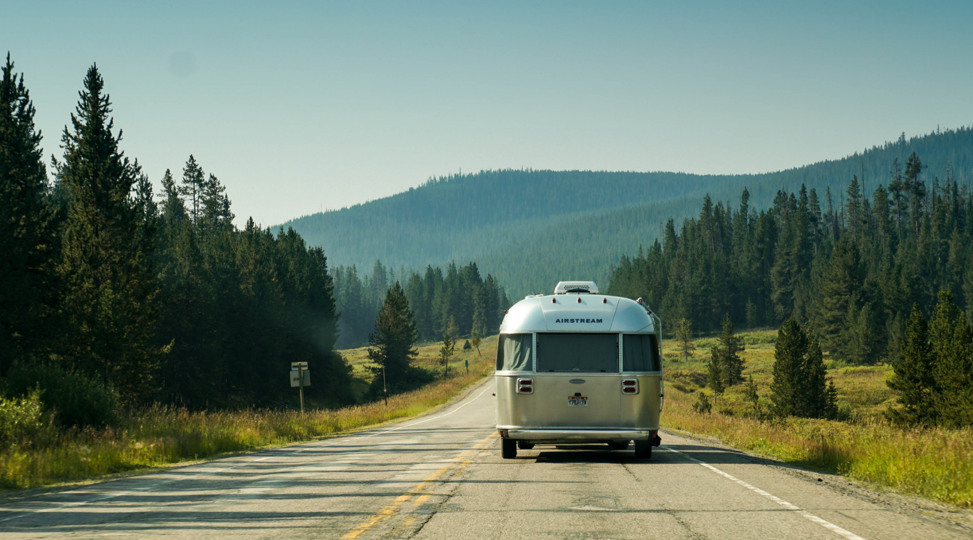 An Airstream trailer is traveling on a scenic road surrounded by dense forests and hills under a clear sky.