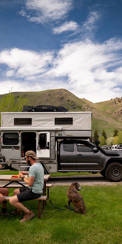 Two people are sitting at a picnic table near a camper truck with a dog sitting beside them, set against a scenic backdrop of mountains and houses.
