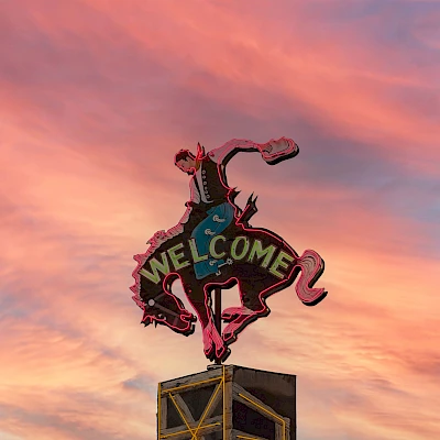 The image shows a sign featuring a cowboy on a bucking horse with the word "WELCOME" against a background of a colorful sunset.