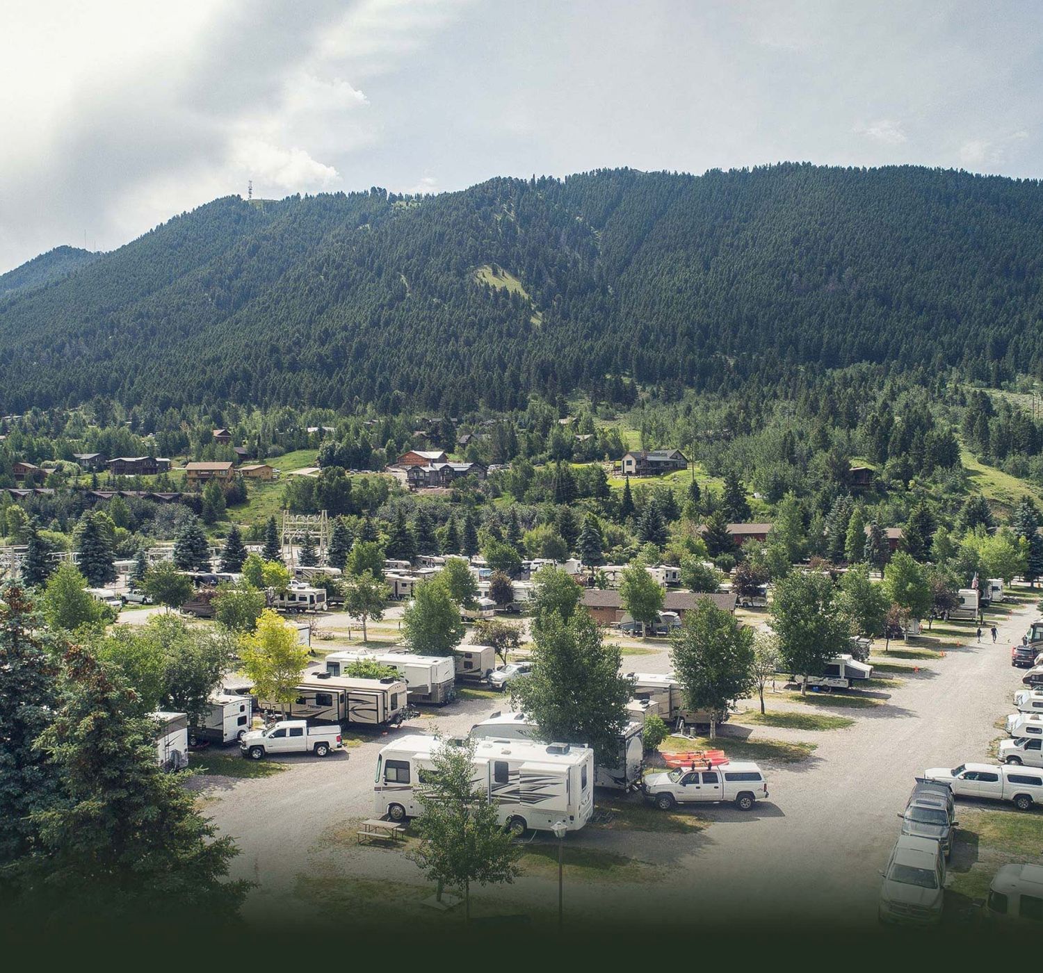 The image depicts a campground with numerous RVs parked among trees, set against a backdrop of forested hills and mountains under a cloudy sky.