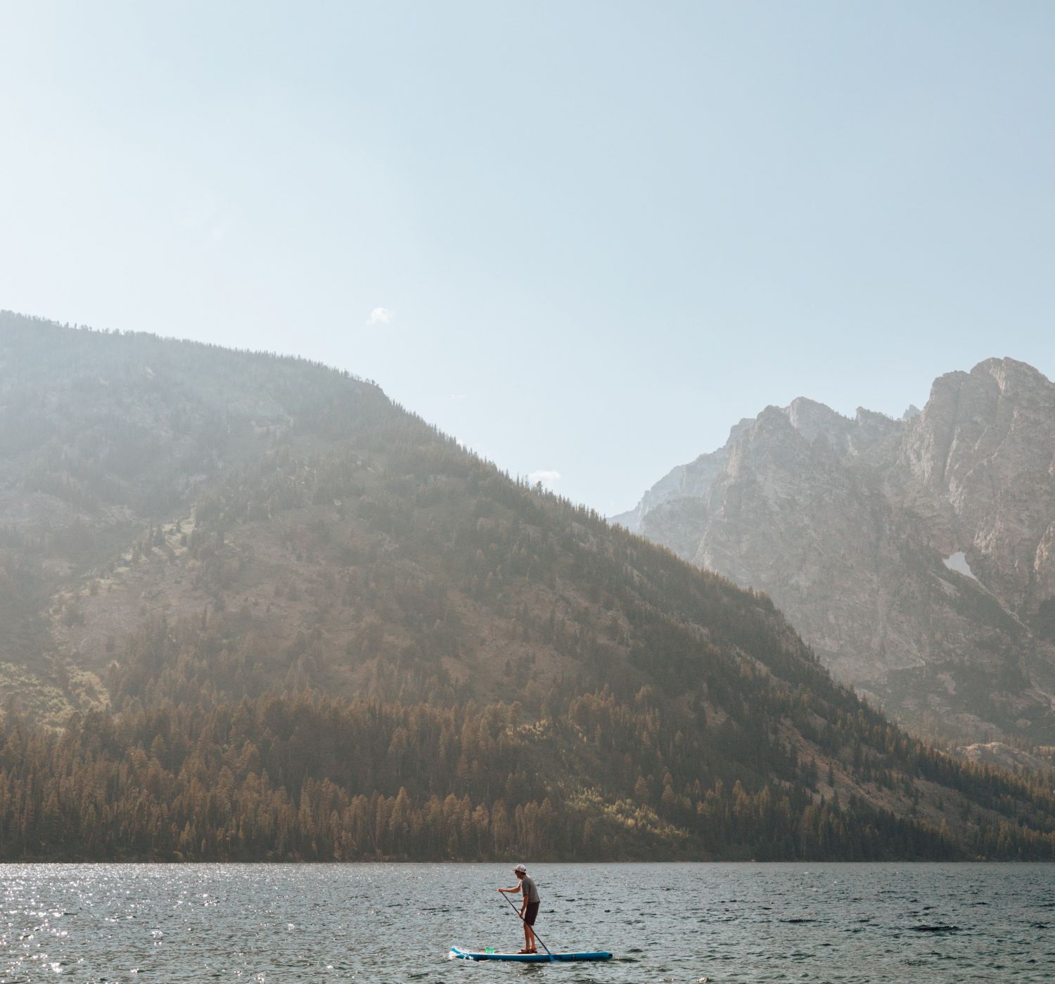 A person is paddleboarding on a lake surrounded by mountains and trees under a clear sky.
