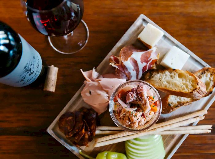 A wooden table with a charcuterie platter, featuring meats, cheeses, bread, and a glass of red wine alongside a bottle of wine and a cork.