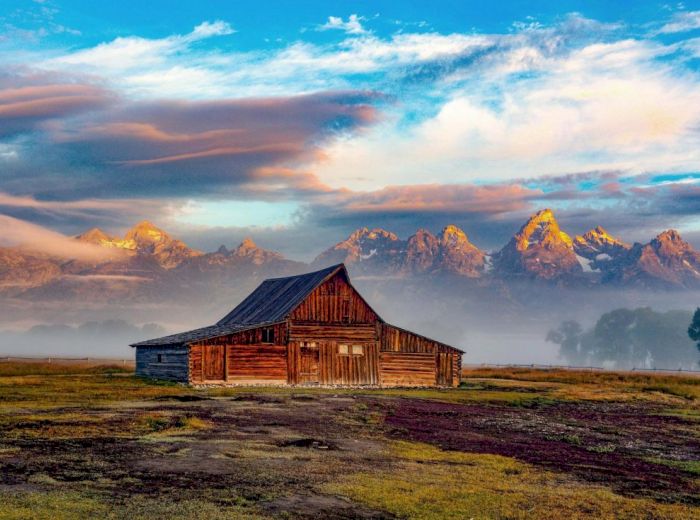 An old wooden barn sits in an open field with a backdrop of misty mountains and a colorful, cloudy sky at sunrise or sunset.