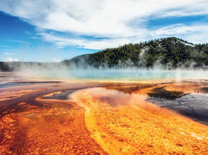 A colorful geothermal hot spring with vibrant orange and yellow bacterial mats, steaming water, and a backdrop of forested hills and a partly cloudy sky.