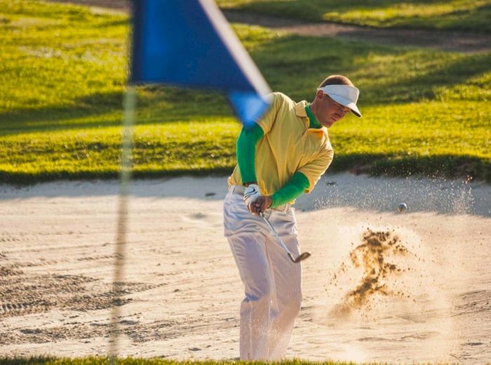 A golfer in yellow shirt and white hat is hitting a shot from a sand bunker on a sunny golf course.