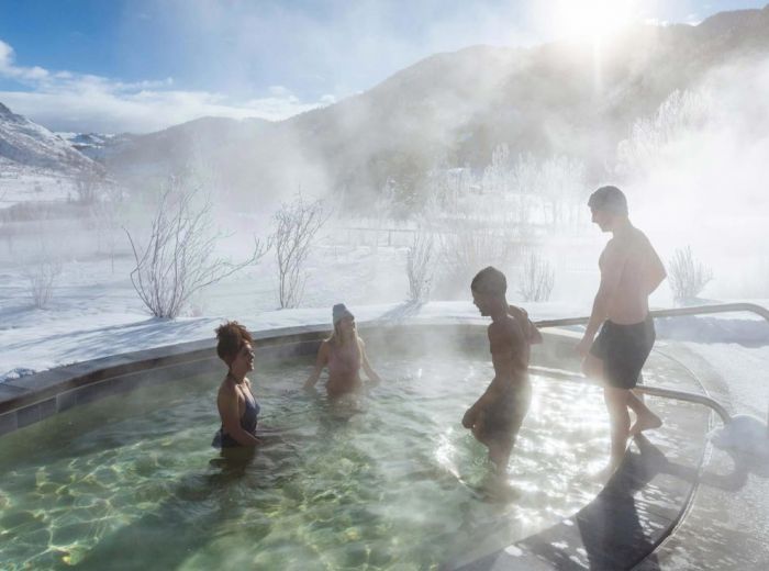 Four people are enjoying a hot spring in a snowy outdoor setting with mountains and a foggy atmosphere due to steam from the warm water.