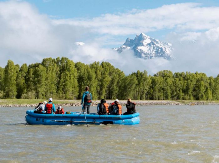 A group of people in life jackets are rafting on a river surrounded by trees, with a snow-capped mountain in the background, under a partly cloudy sky.