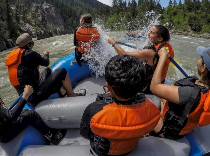 A group of people wearing life jackets are whitewater rafting on a river in a forested area. One person is getting splashed with water.