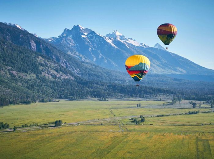Two colorful hot air balloons float over a scenic valley with mountains in the background under a clear blue sky.