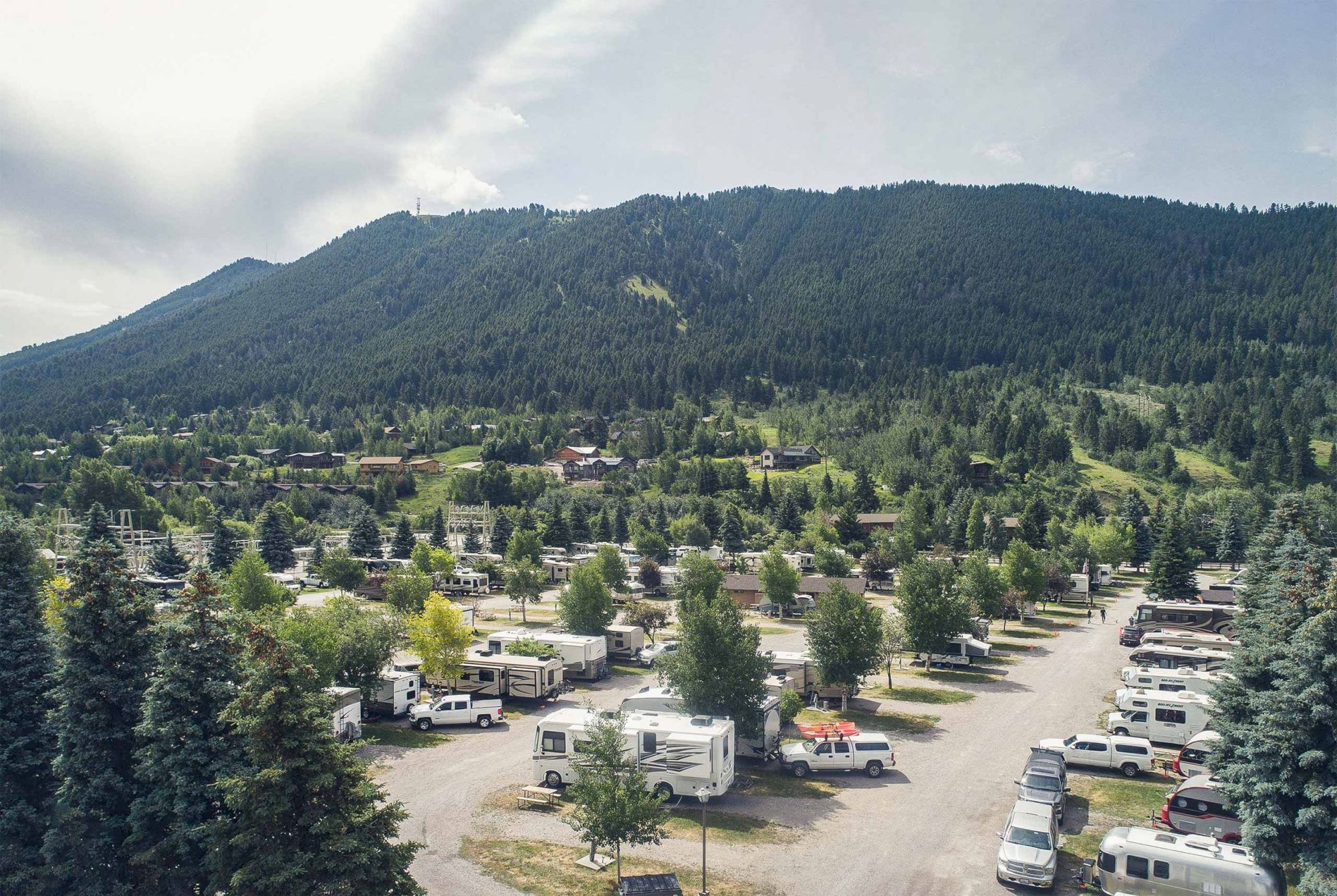 The image shows a campground with numerous RVs situated among trees, set against a backdrop of forested mountains.
