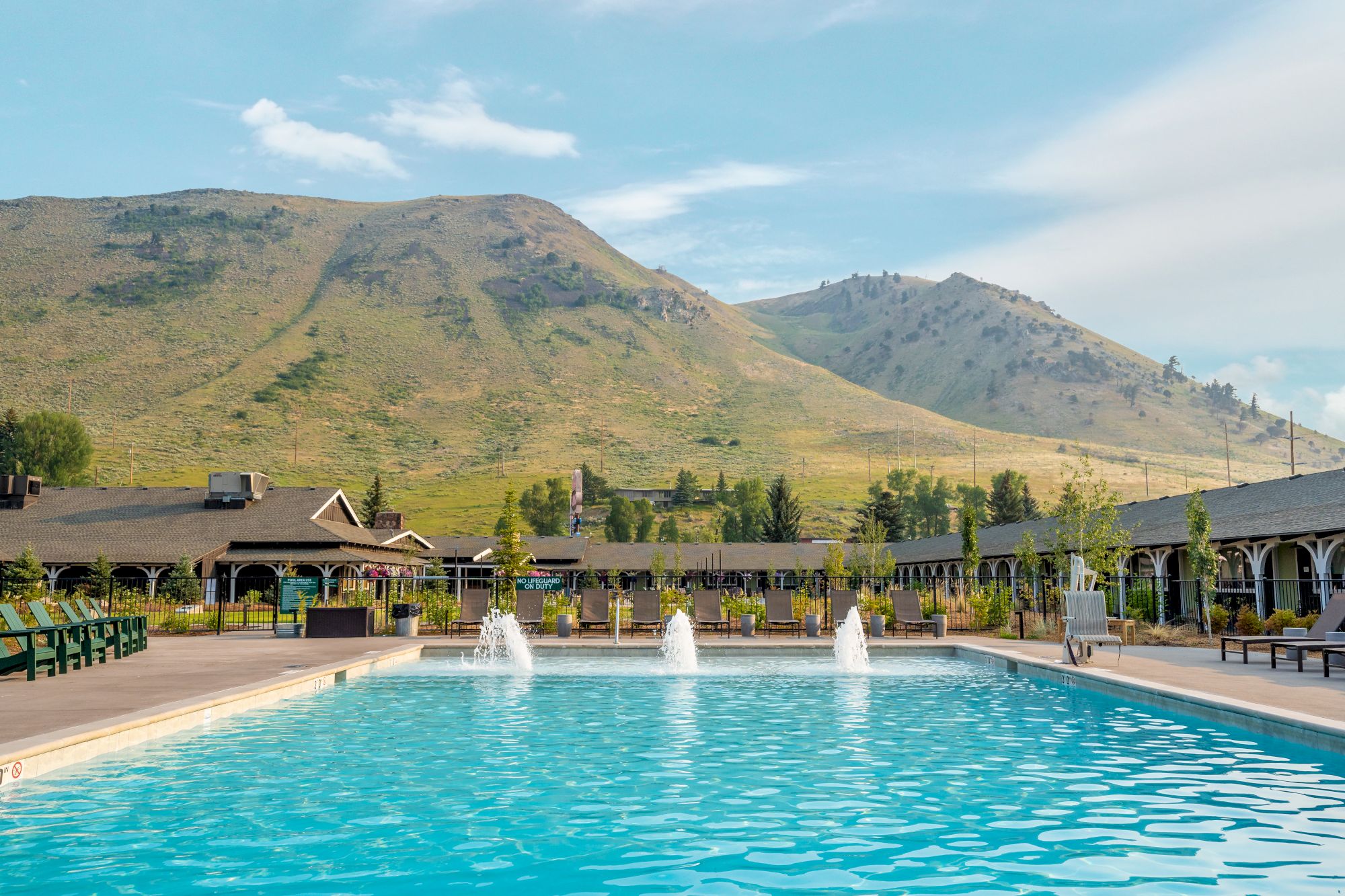 An outdoor pool with fountains in the foreground, set against a backdrop of mountains and clear skies with buildings surrounding it.
