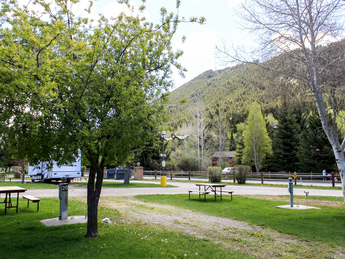 The image shows a serene campsite with picnic tables, grassy areas, and trees, set against a backdrop of hills and mountains.