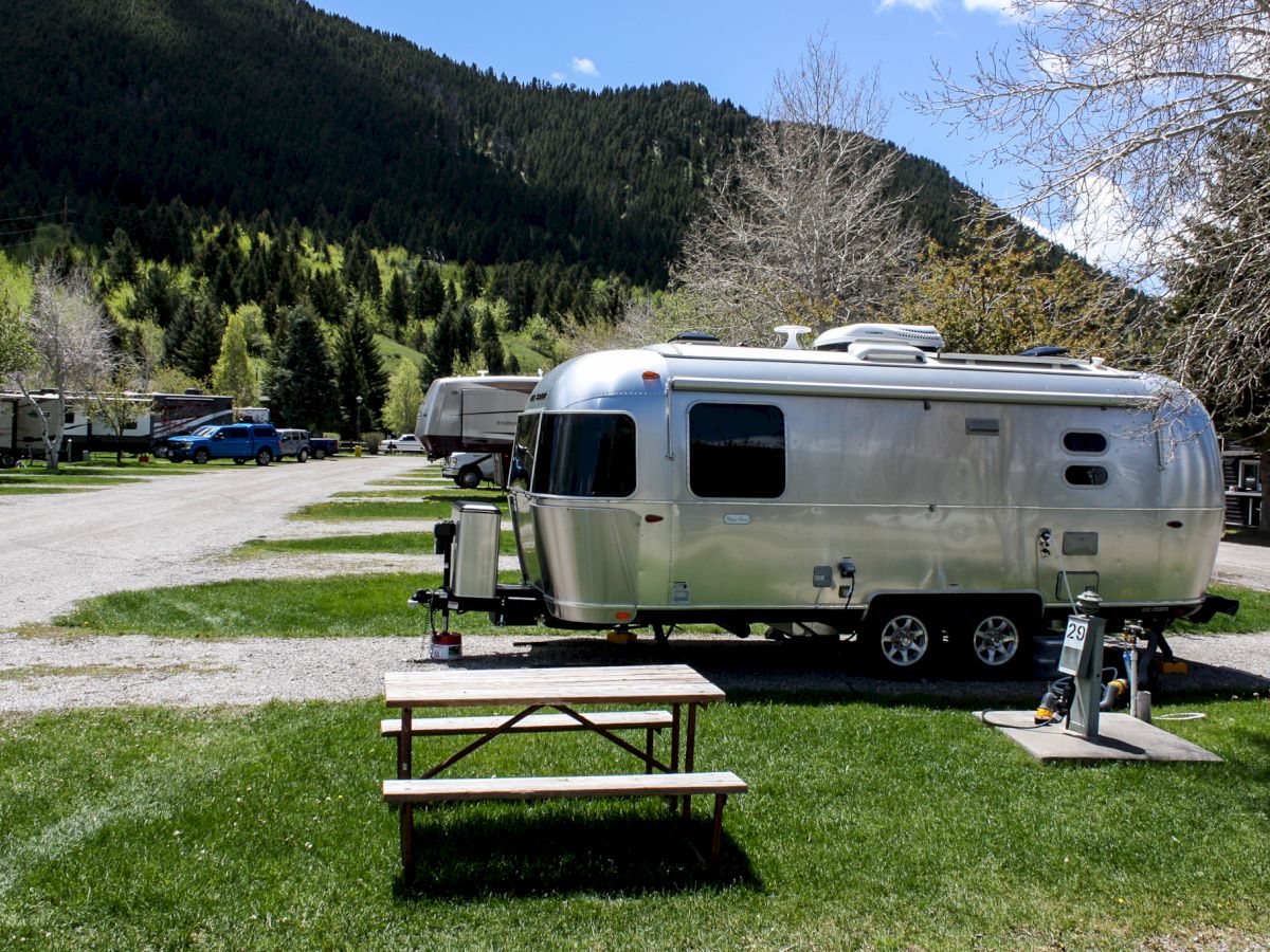 The image shows a shiny, modern Airstream trailer on a grassy campsite with a picnic table, surrounded by trees and mountains under a blue sky.