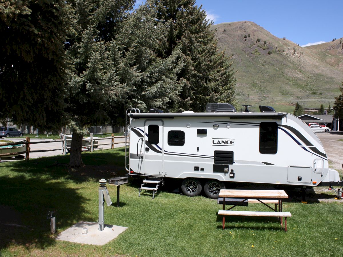 The image shows a Lance trailer parked on a grassy area with a picnic table and trees, against a backdrop of mountains and a clear blue sky.