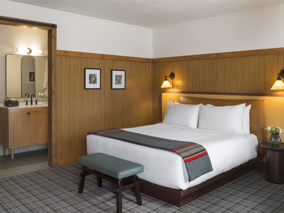 A cozy hotel room features a neatly made bed, wooden paneling, bedside lamps, a small bench, and an open door leading to a bathroom.