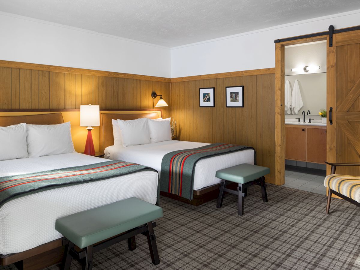 The image shows a cozy hotel room with two beds, plaid carpeting, wooden accents, and a sliding barn door leading to a bathroom.