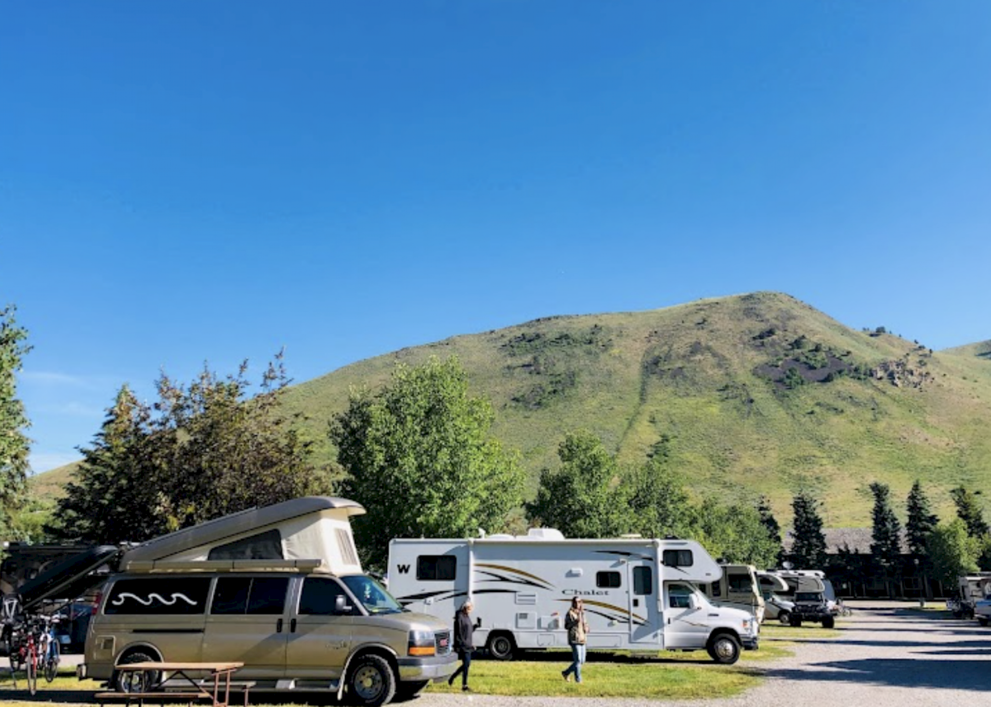 Several RVs are parked in a camping area with a background of green hills under a clear blue sky.