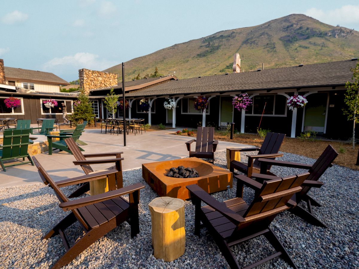 An outdoor seating area with wooden chairs around a fire pit, overlooked by a building and mountains in the background.