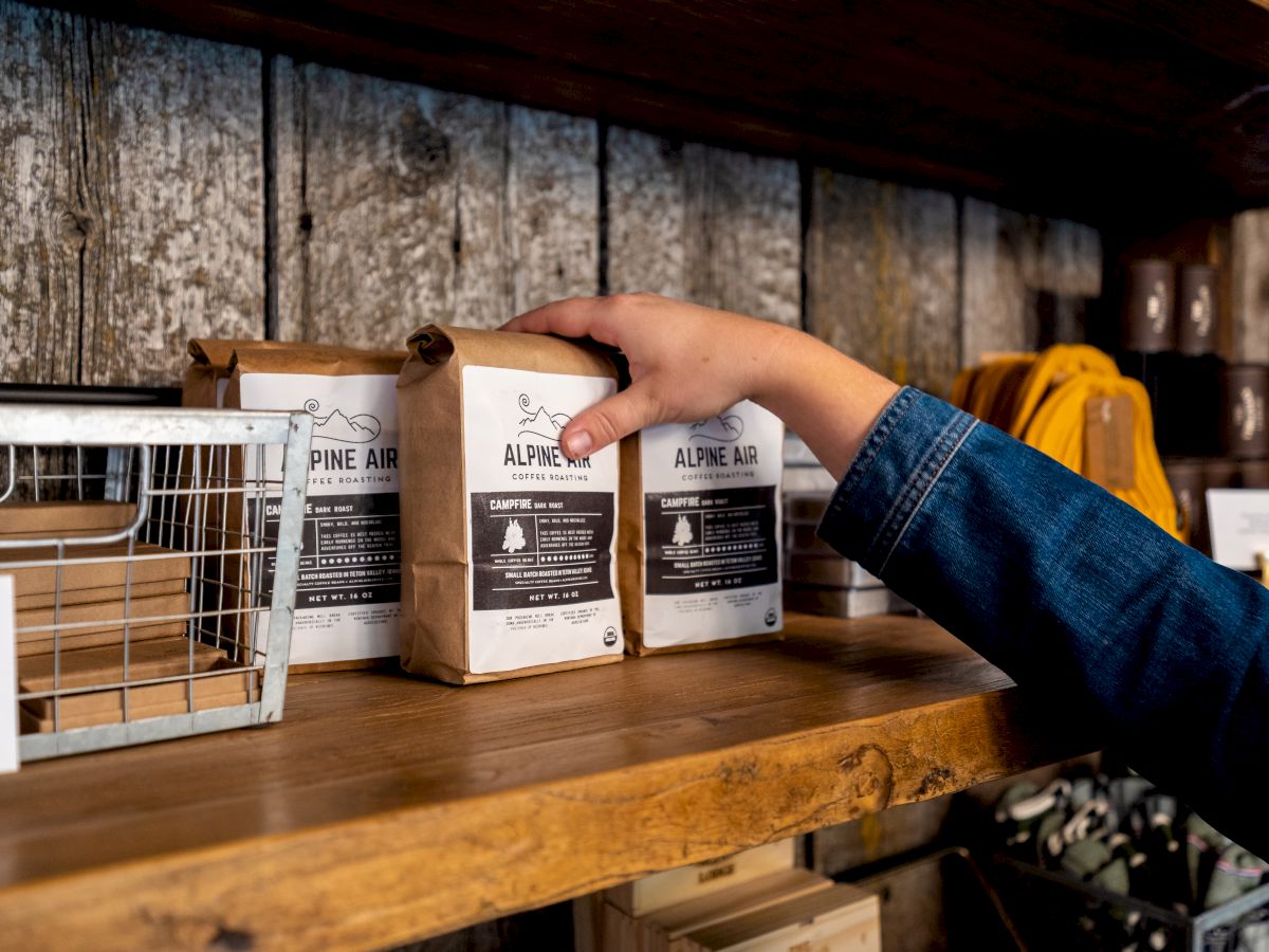 A person’s hand reaching for a bag among neatly arranged packs on a rustic wooden shelf, with various items beside it in a cozy setting.